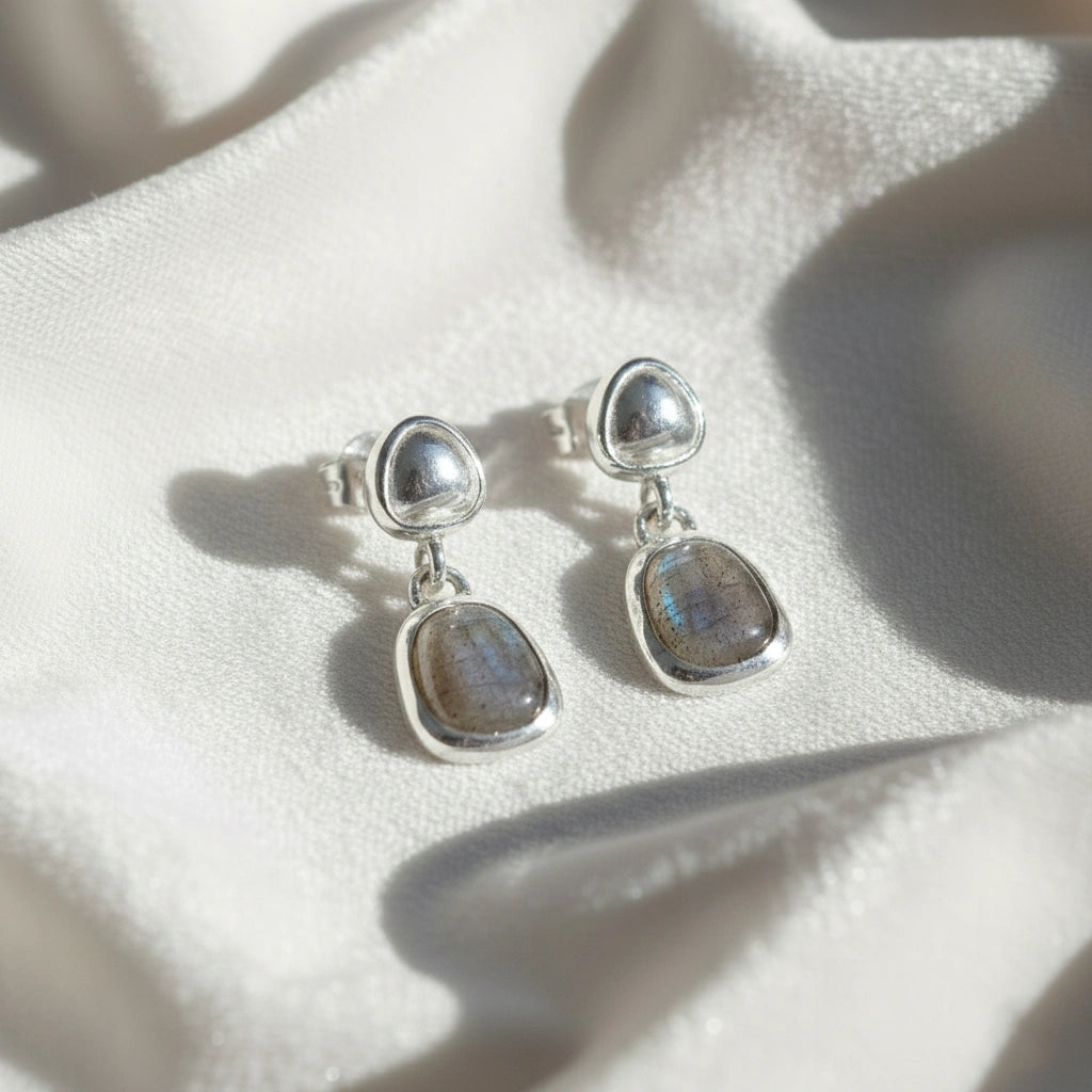 Tiny earrings featuring silver studs with labradorite charm stones angled downward on a soft white fabric