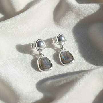 Tiny earrings featuring silver studs with labradorite charm stones angled downward on a soft white fabric