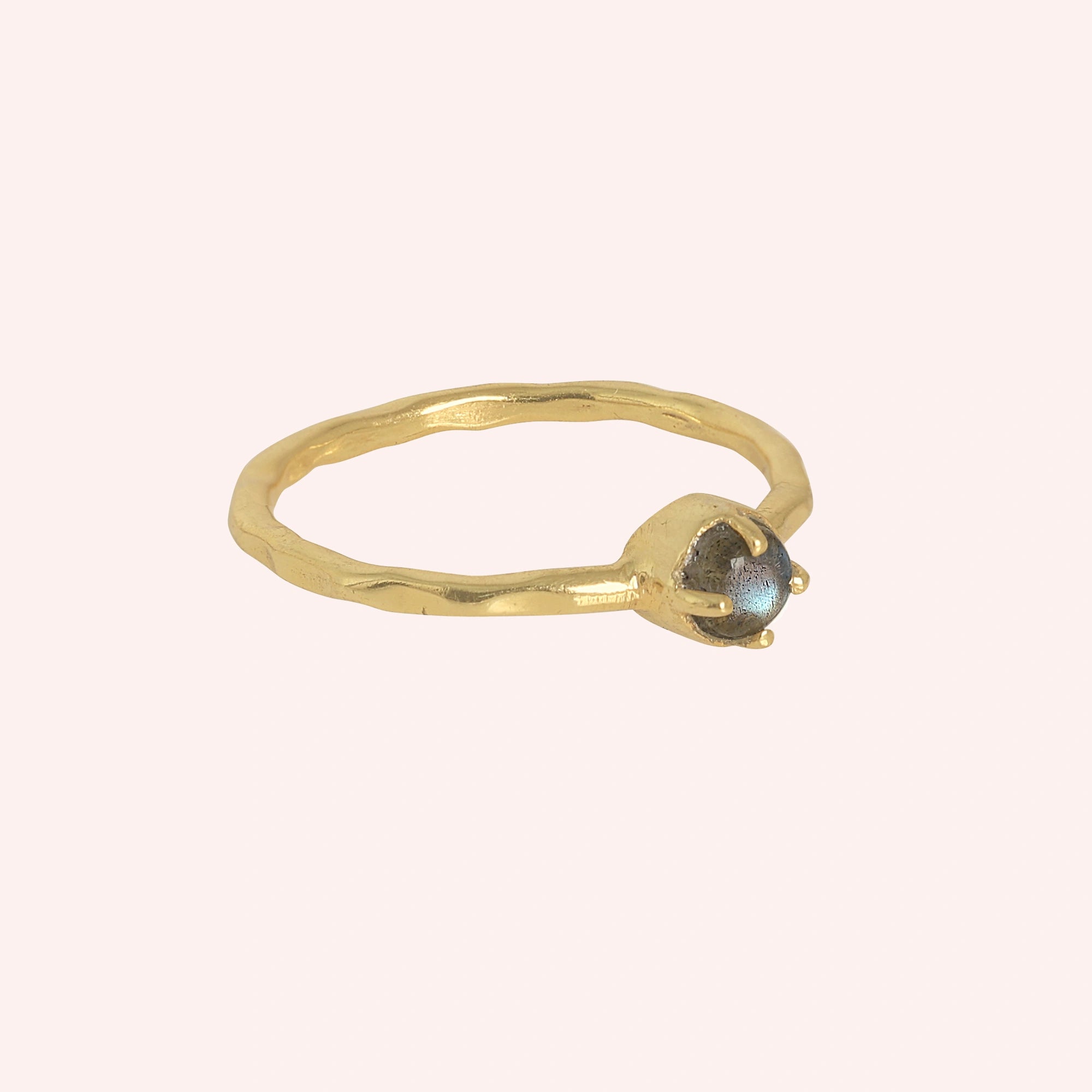 Minimalist ring featuring a small round labradorite stone in a gold prong setting on a light pink background at a slight right angle