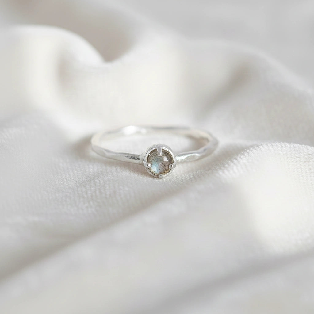 Minimalist ring featuring a round labradorite stone in a silver prong setting on a white fabric surface straight-on angle