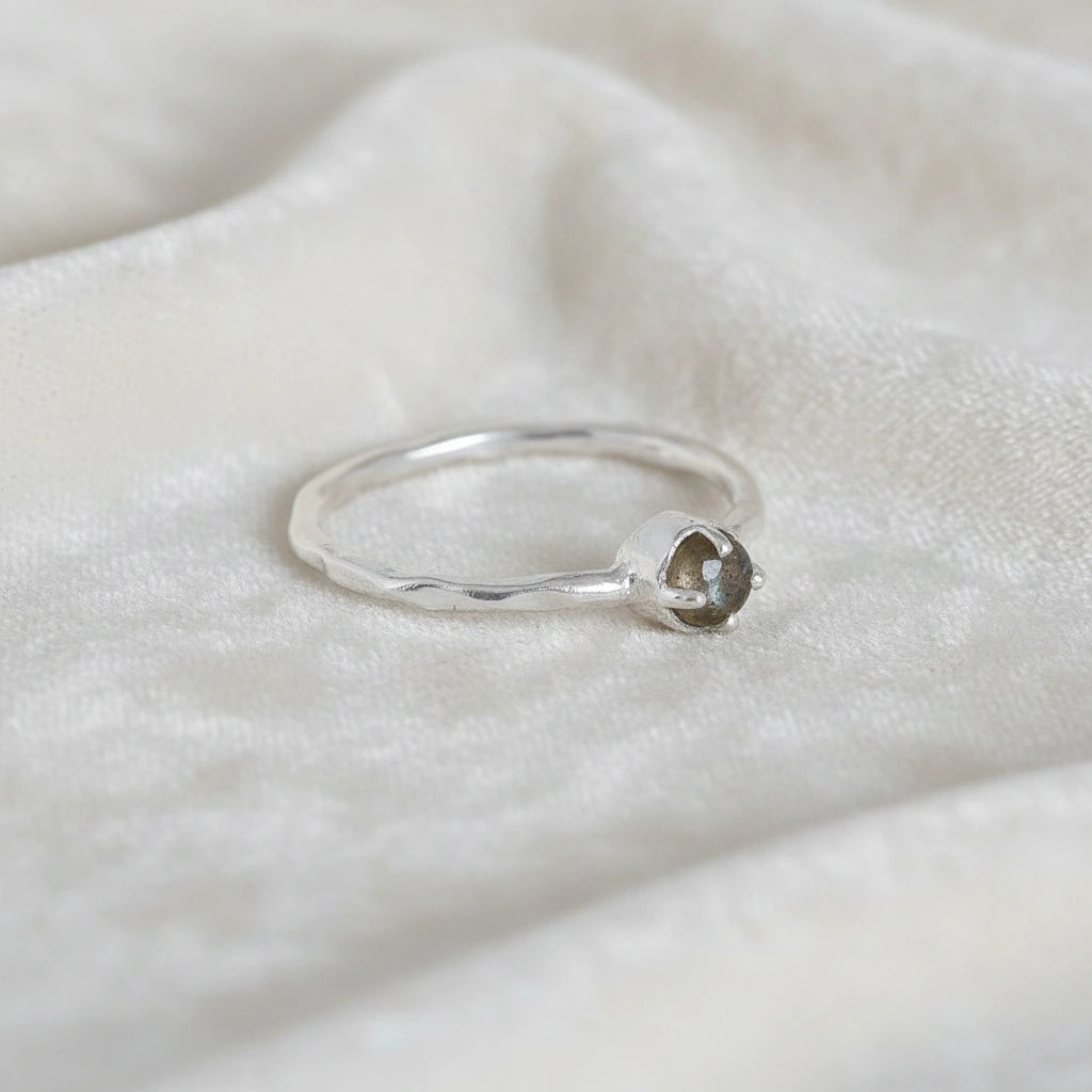 Minimalist ring featuring a small round labradorite stone in a silver prong setting on a white fabric at a slight right angle