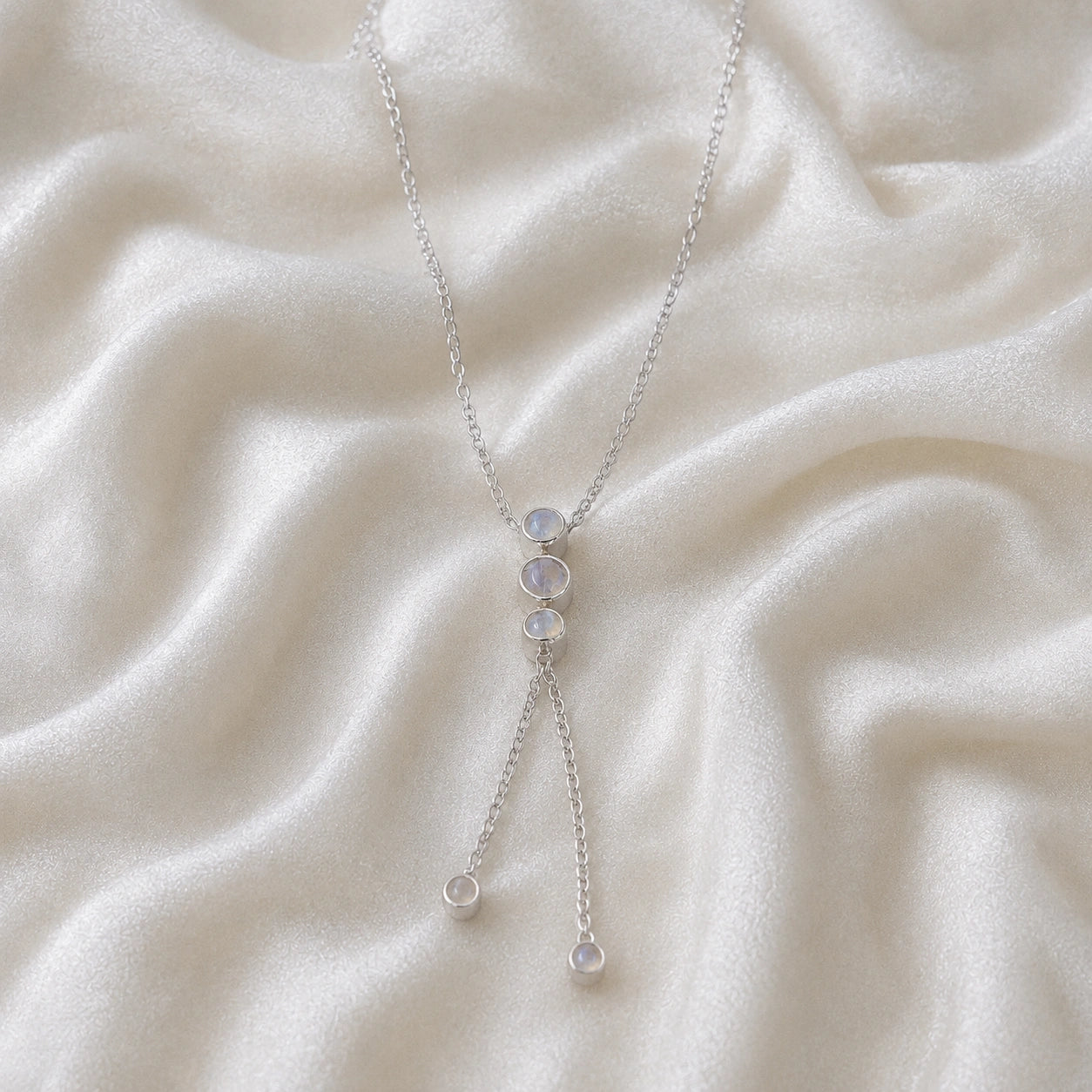 Lariat_necklace_in_sterling_silver_at_front_angle_featuring_moonstone_drops_on_soft_fabric_background