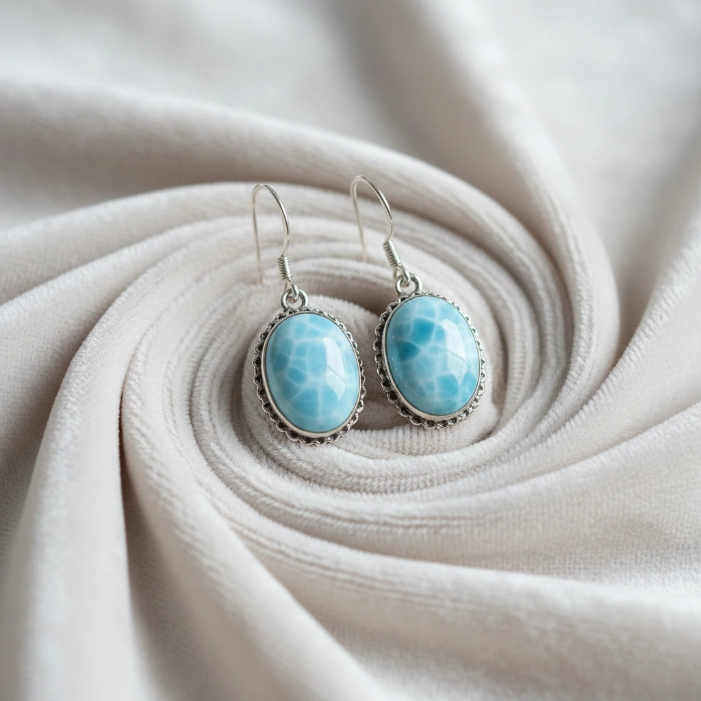 Silver dangling earring featuring oval blue larimar stones on a pleated off white velvet background side-top angle