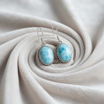 Silver dangling earring featuring oval blue larimar stones on a pleated off white velvet background side-top angle