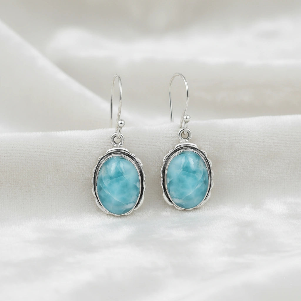 Silver dangling earring blue Larimar oval stone set in silver bezel viewed from a straight-on angle on off white velvet cloth