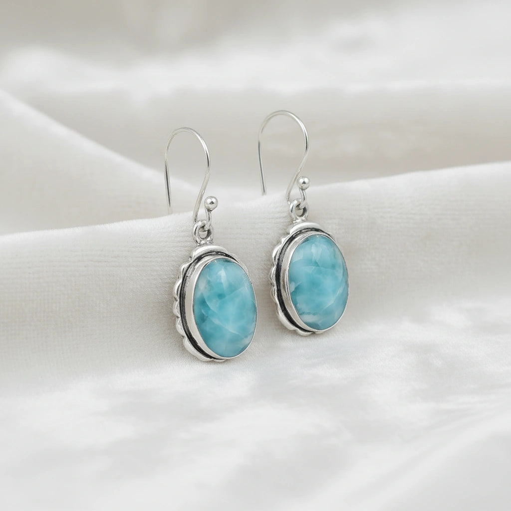 Silver dangling earring blue Larimar oval gemstone set in bezel detail close up angle on an off white velvet cloth background