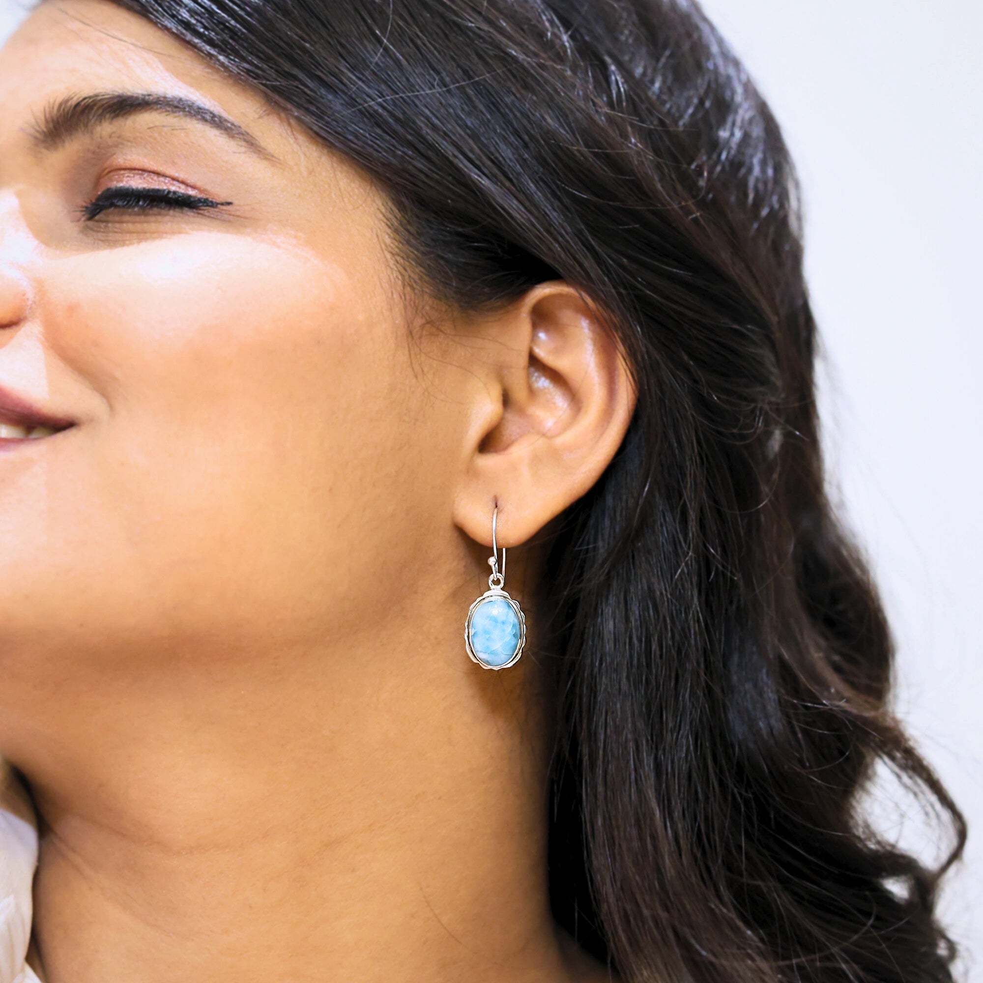 Silver dangling earring blue Larimar oval stone set in serrated bezel worn by a smiling model close up side profile angle