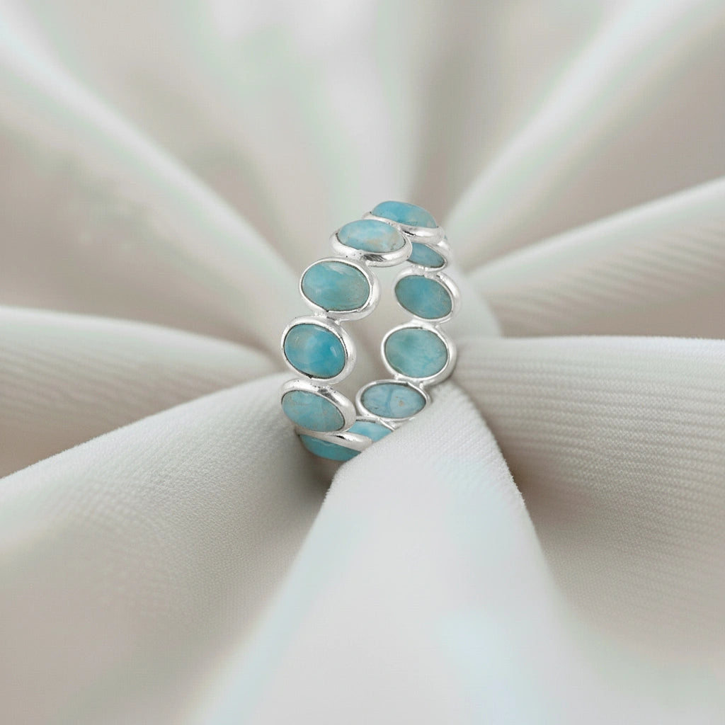 Silver Larimar eternity ring with oval blue stones standing upright on a softly draped creamy white velvet cloth.