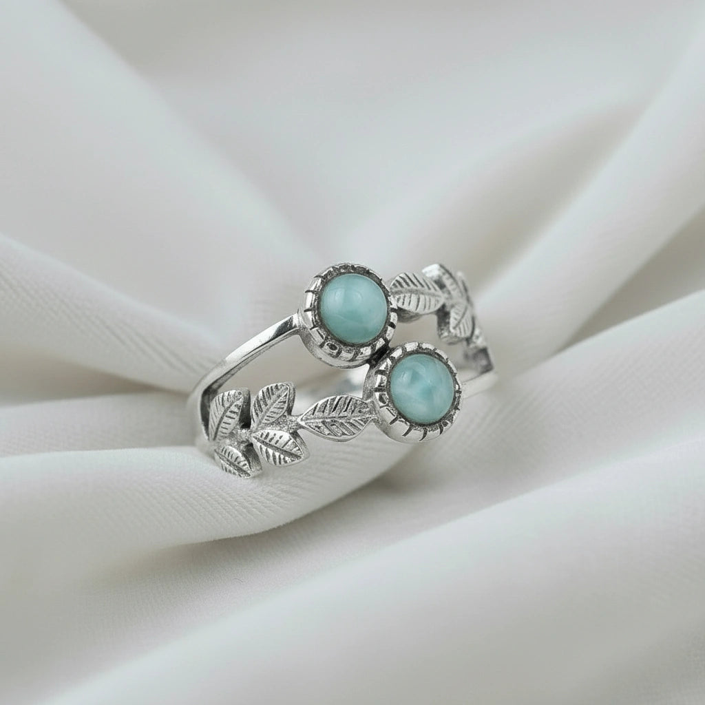 Detailed sterling silver leaf ring with two round Larimar stones on a white draped fabric background viewed at an angle