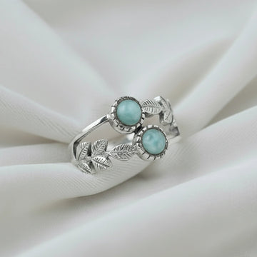 Detailed sterling silver leaf ring with two round Larimar stones on a white draped fabric background viewed at an angle