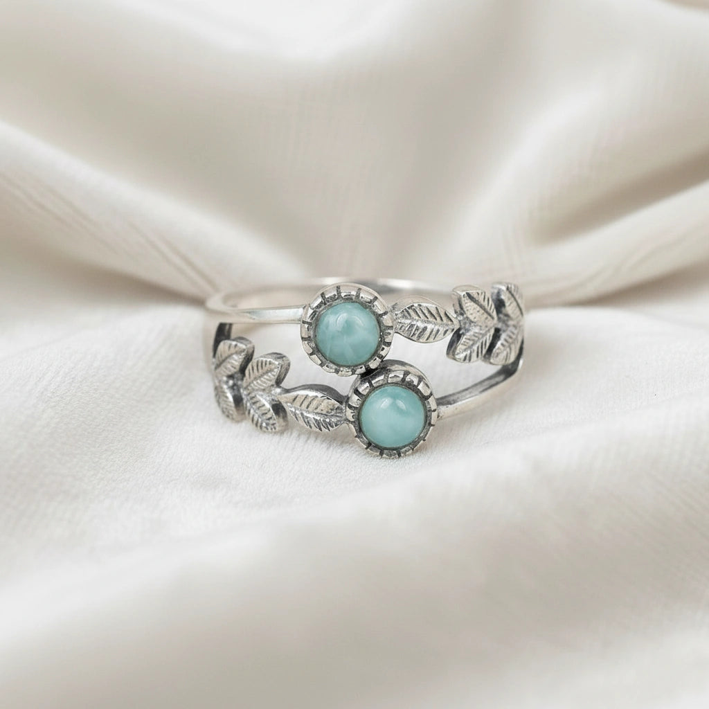 Detailed sterling silver leaf ring with two round Larimar stones on a white velvet fabric background viewed straight on