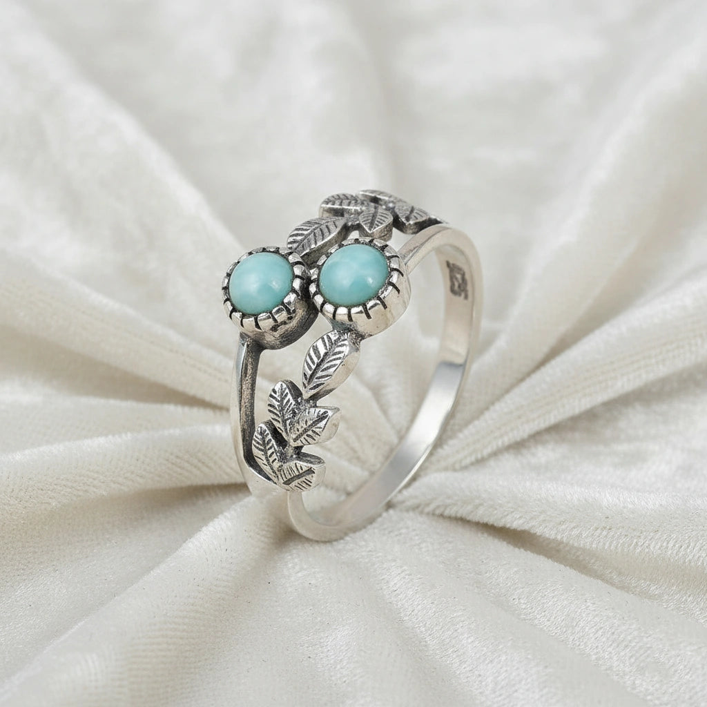 Detailed sterling silver leaf ring with two round Larimar stones on a white draped fabric background viewed from an angle