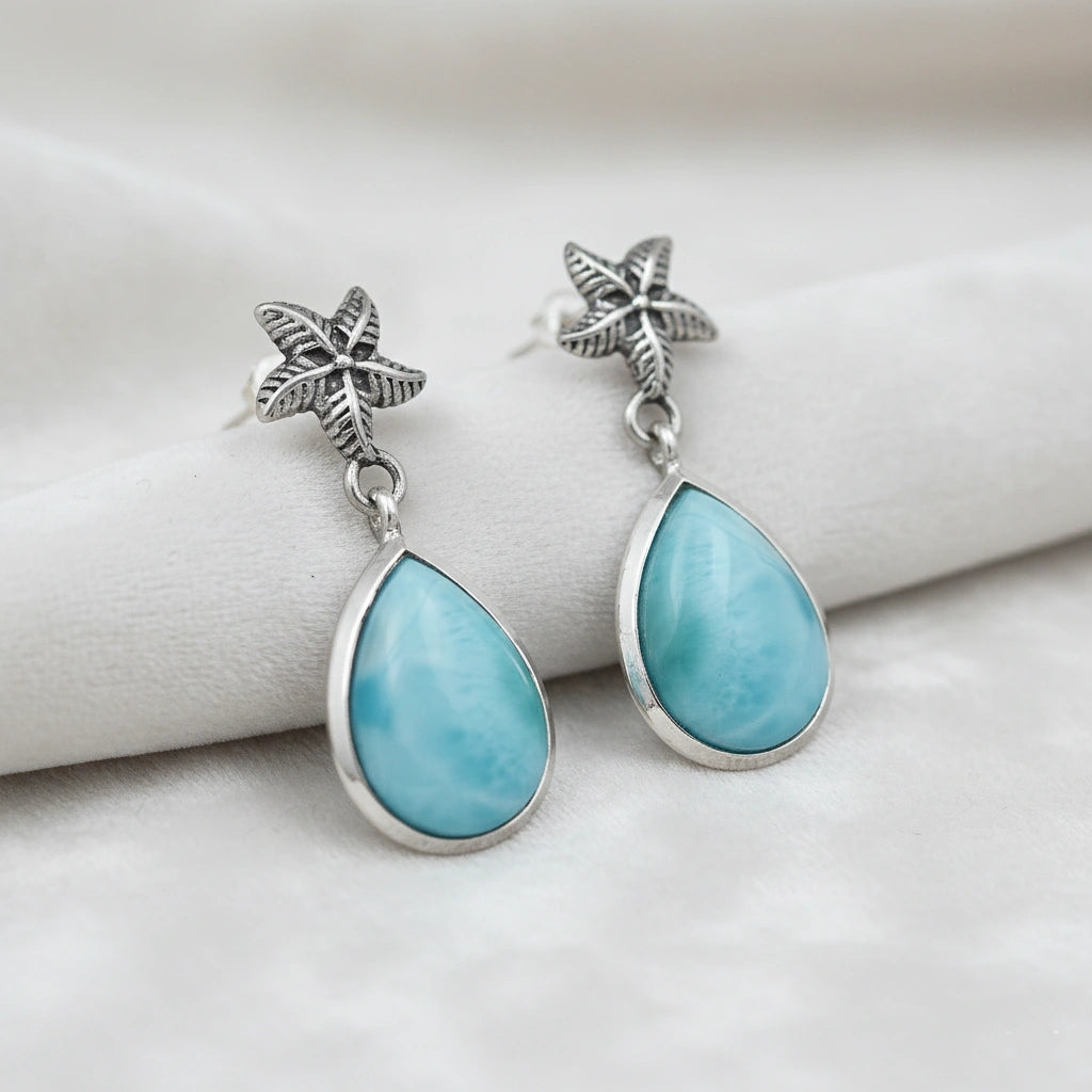 Starfish earrings featuring blue Larimar stones drop down on a white velvet surface slightly angled close up studio photo