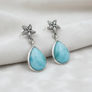Starfish earrings featuring blue Larimar stones drop down on a white velvet surface slightly angled close up studio photo