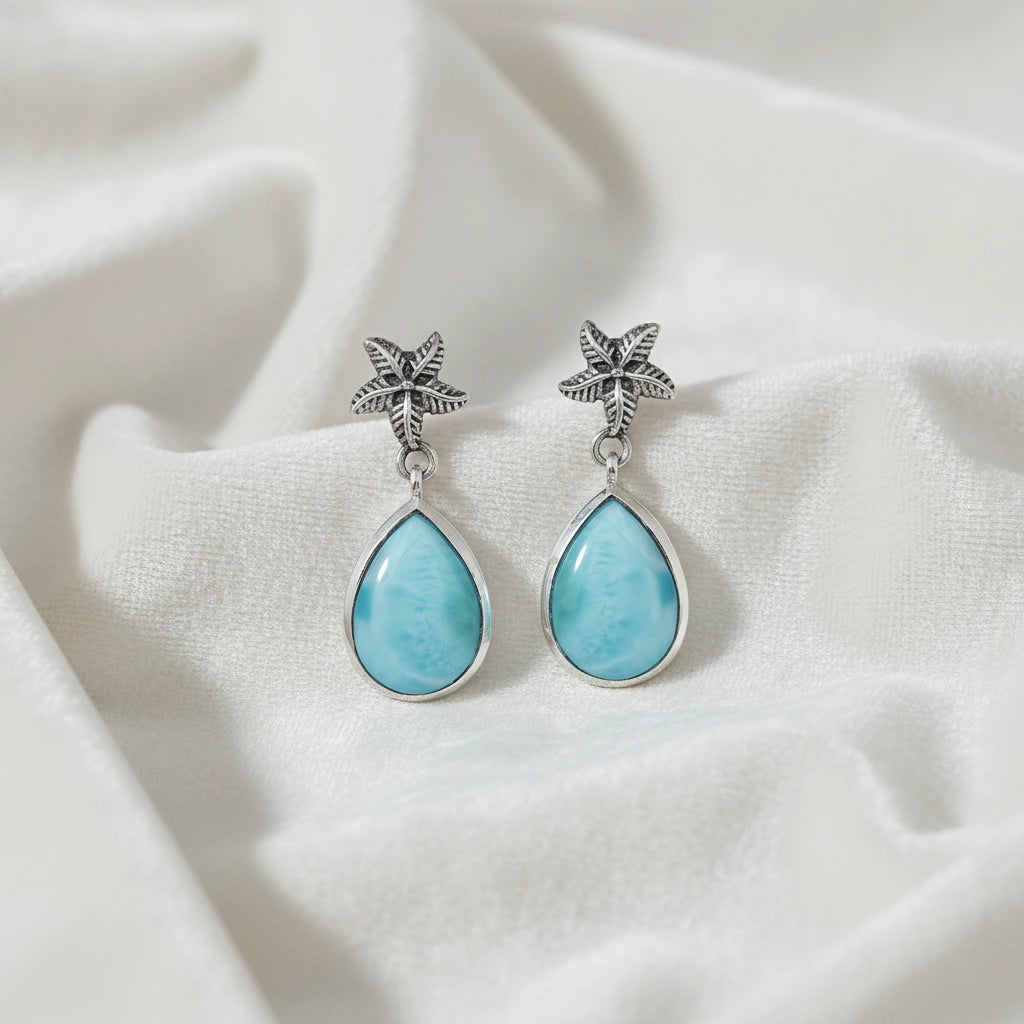Starfish earrings with blue Larimar teardrop gems lie on a white draped velvet fabric taken straight on with a slight elevation