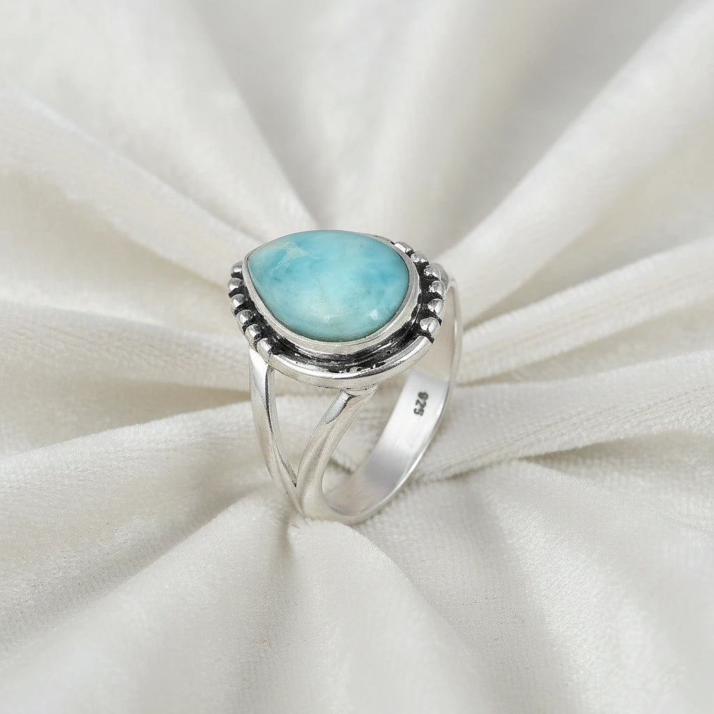 Detailed silver teardrop ring with a Larimar stone resting on a white draped fabric background viewed from an elevated angle