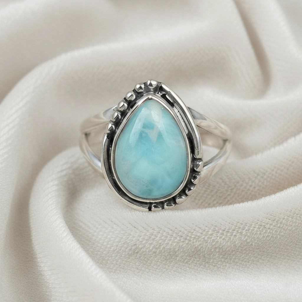 Detailed silver teardrop ring with a Larimar stone resting on an off white draped velvet fabric background viewed straight on