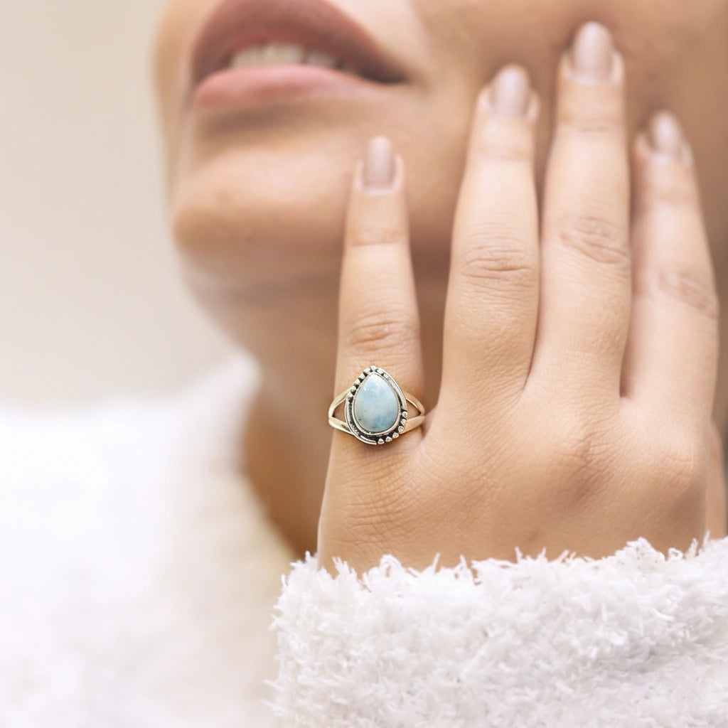 Detailed silver teardrop ring with a Larimar stone resting on an off white draped velvet fabric background viewed straight on
