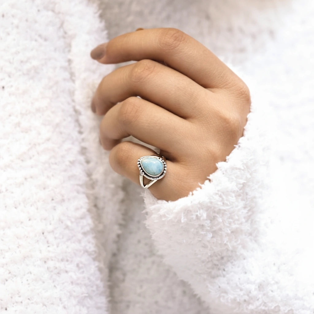 Close up of a silver teardrop ring with a Larimar stone worn on a hand against a white sweater viewed from a slight side angle
