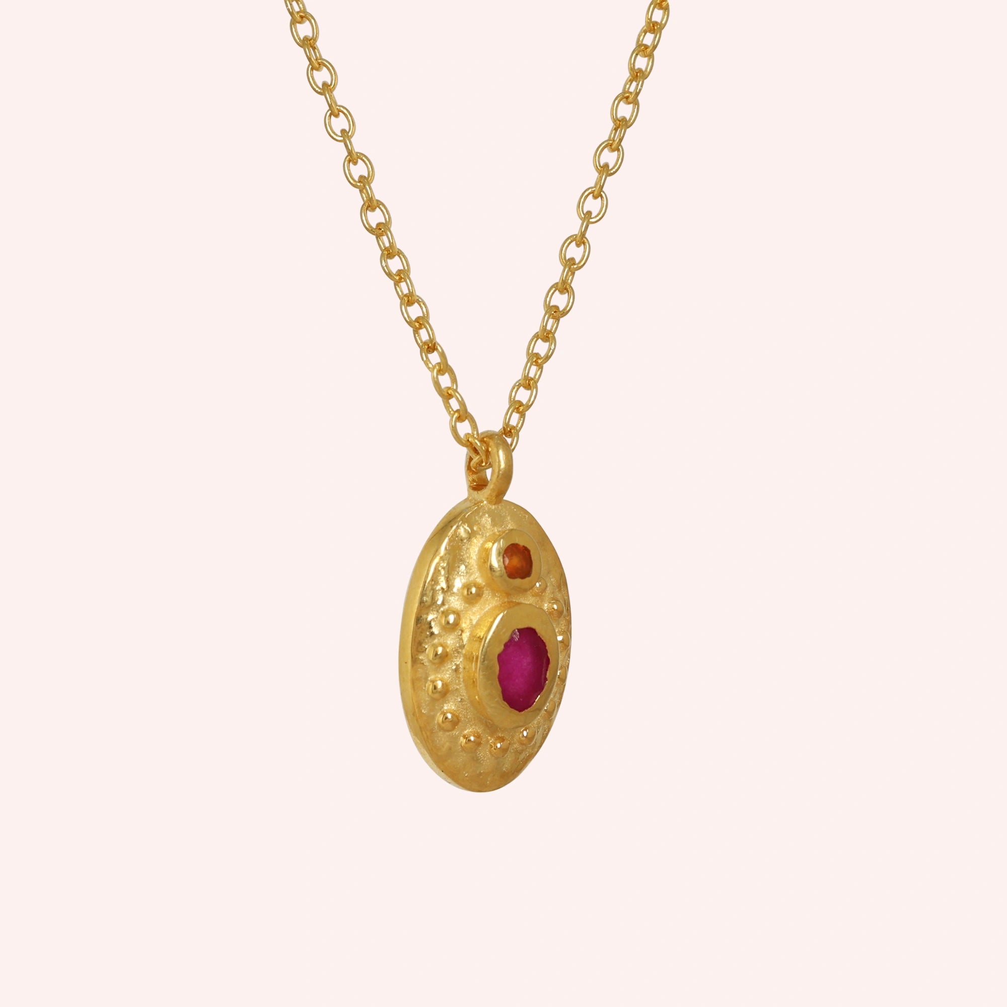 Medallion_necklace_in_gold_plated_silver_slight_angle_with_oval_pendant_with_ruby_and_carnelian_stones_on_solid_pink_background