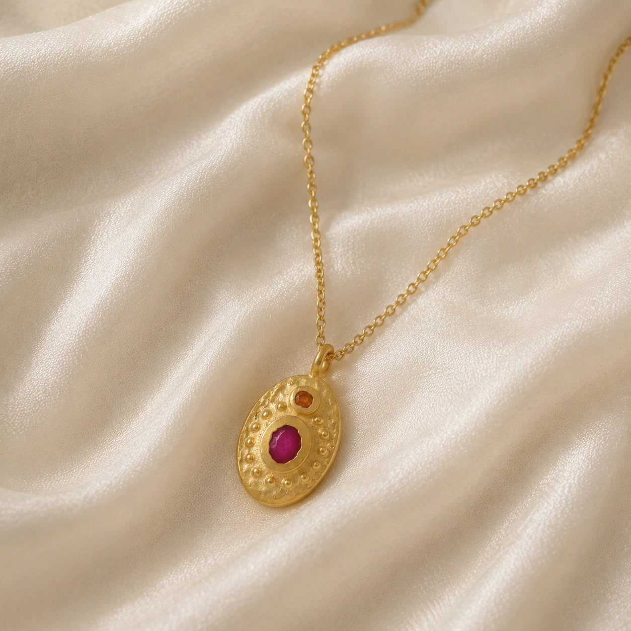 Medallion_necklace_in_gold_plated_silver_slight_angle_with_oval_pendant_with_ruby_and_carnelian_stones_on_soft_fabric_background