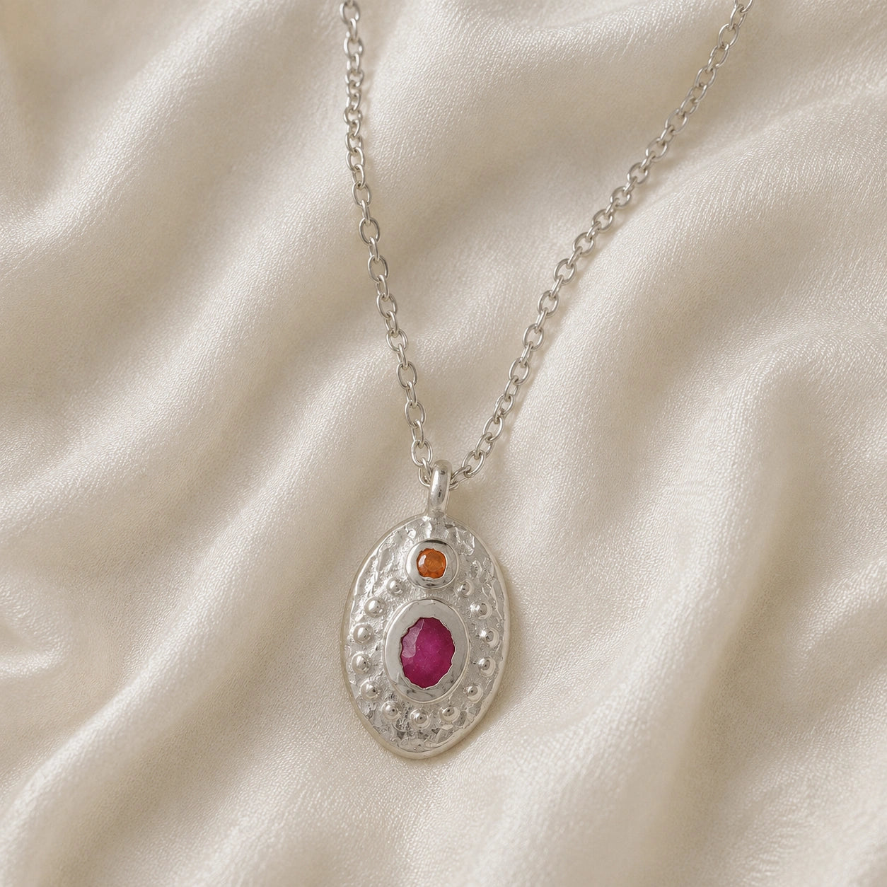Medallion_necklace_in_sterling_silver_front_angle_with_oval_pendant_with_ruby_and_carnelian_stones_on_soft_fabric_background