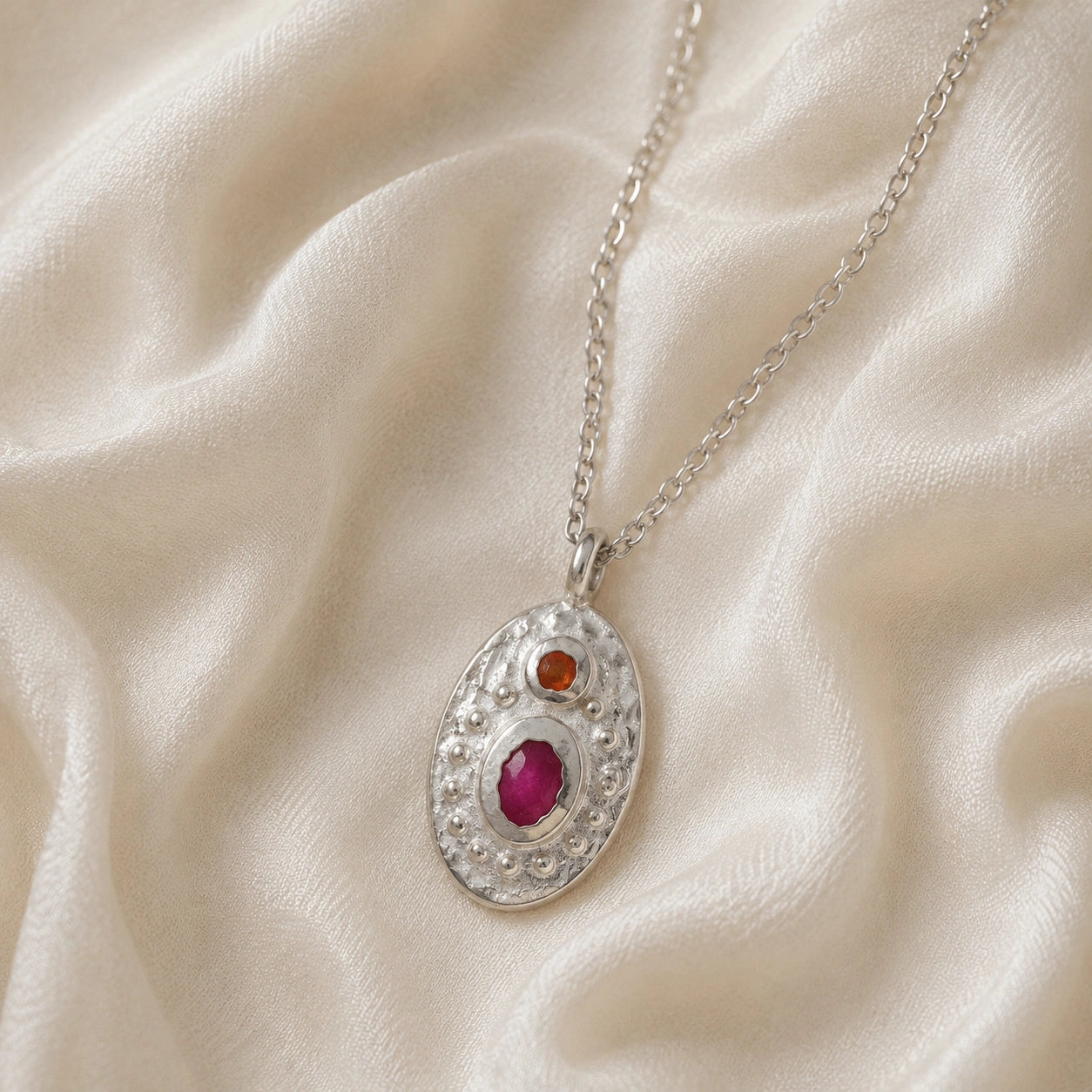Medallion_necklace_in_sterling_silver_slight_angle_with_oval_pendant_with_ruby_and_carnelian_stones_on_soft_fabric_background