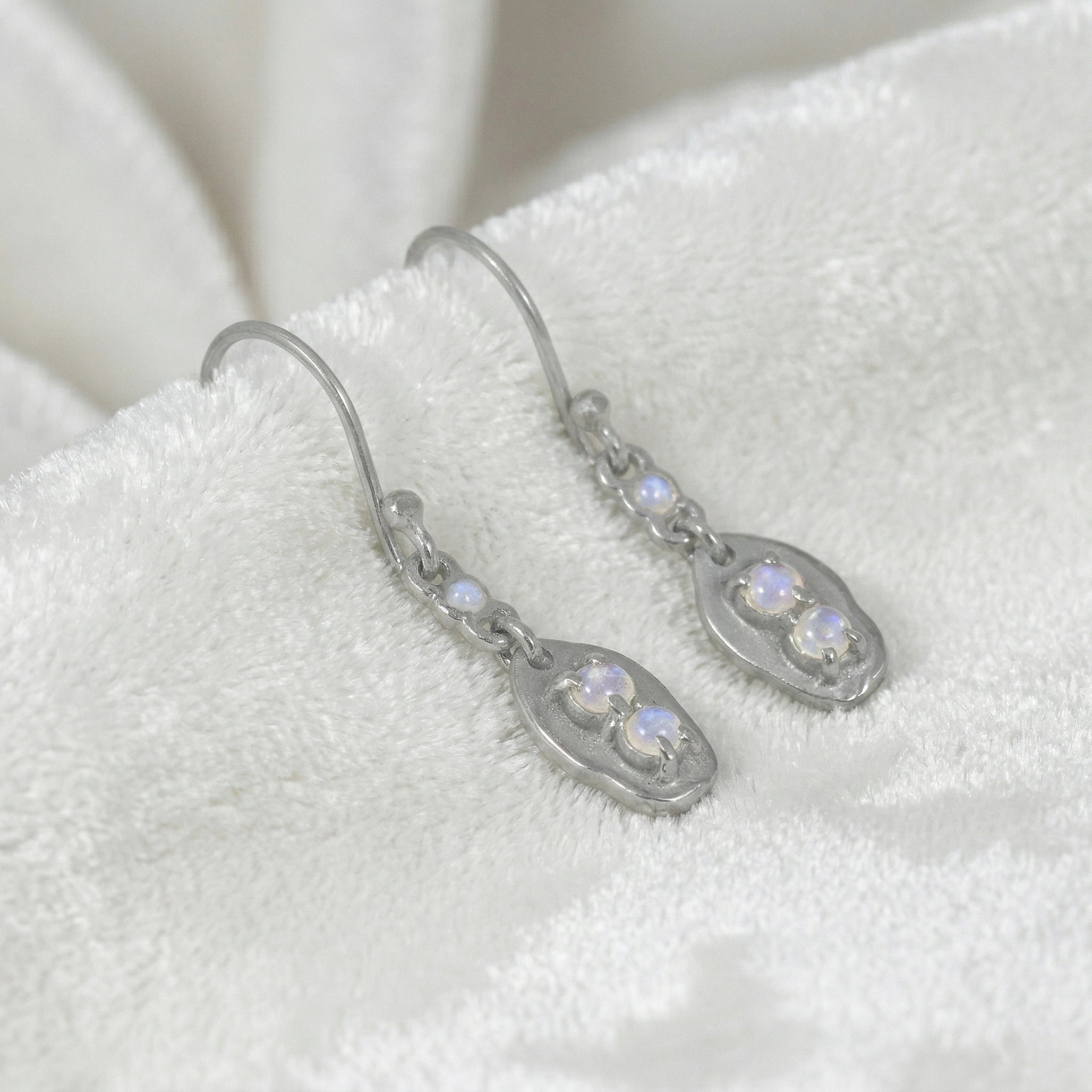 Moonstone_earrings_sterling_silver_drop_design_with_three_moonstones_in_slightly_angled_view_on_soft_white_fabric_background