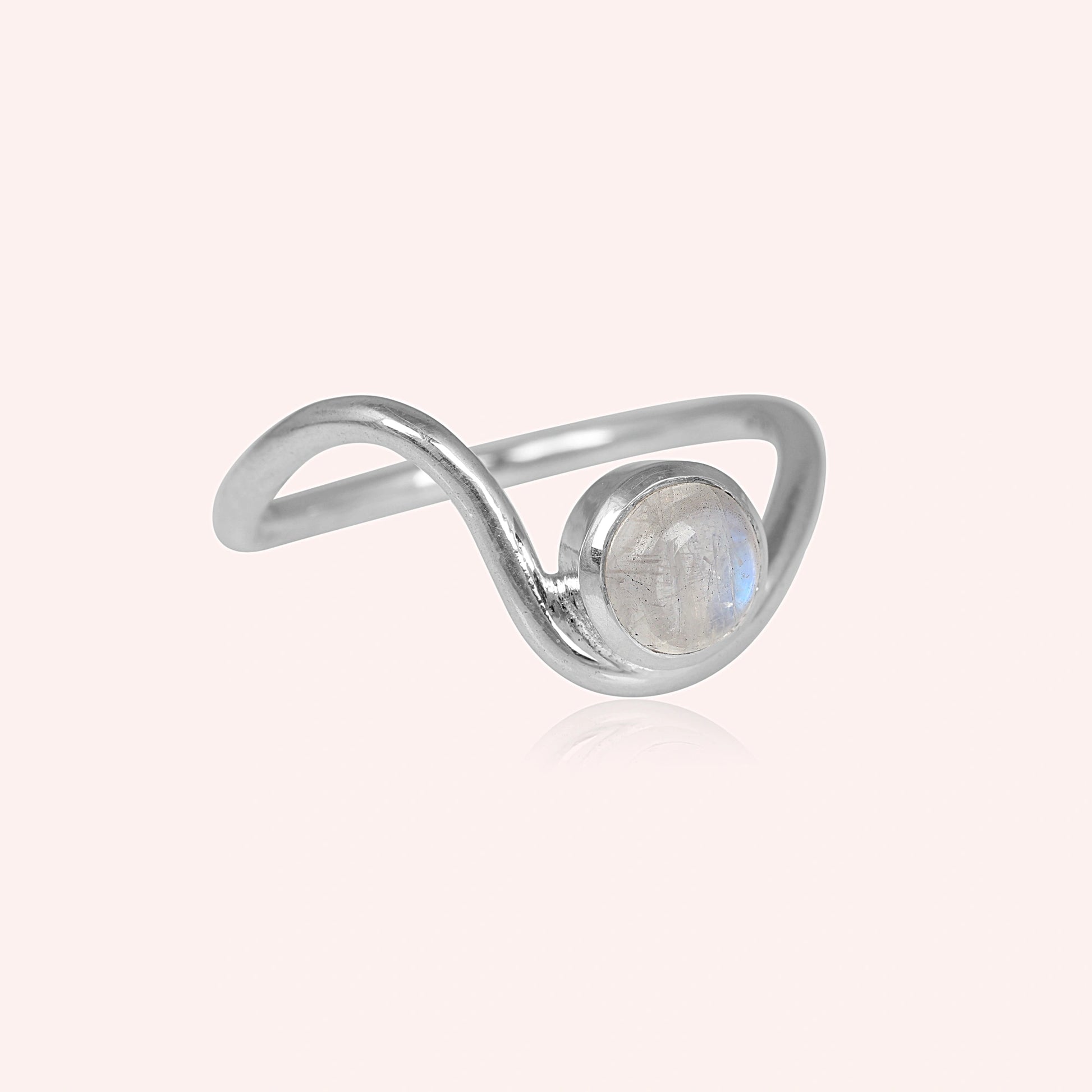 A silver moonstone ring with a round moonstone and a wavy wave ring band at a tilted angle over a light pink plain background