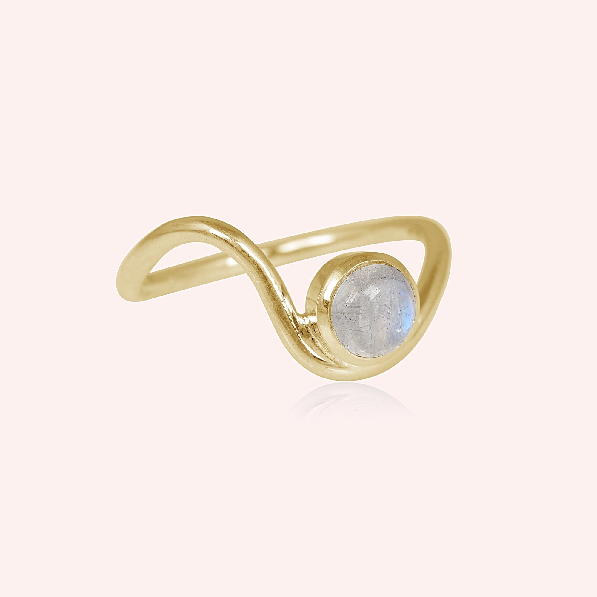 A gold plated moonstone ring featuring a round moonstone and a wave ring band design at a side angle over a light pink background