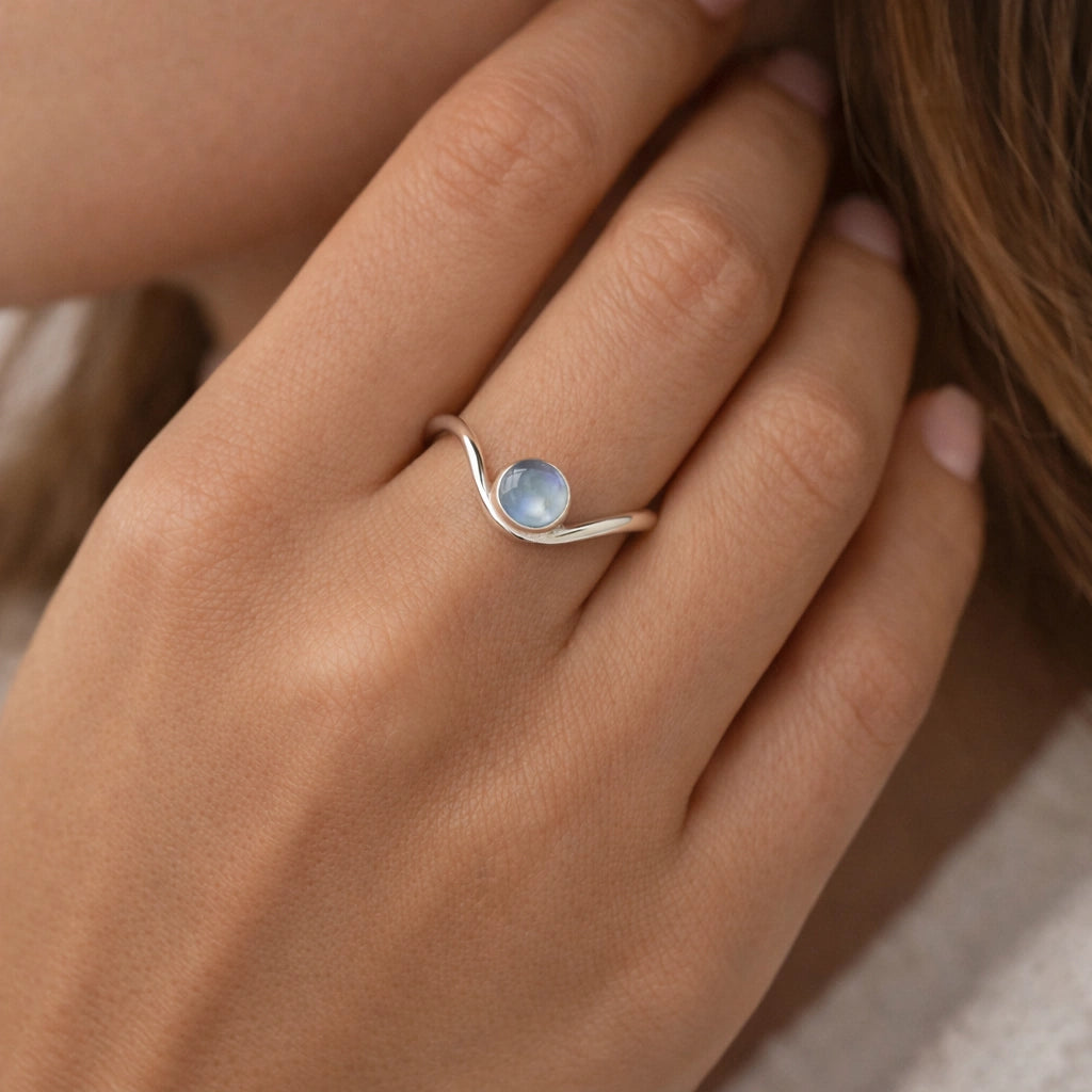 A silver moonstone ring with a round moonstone and a wavy wave ring band at a tilted angle over an off white fabric background