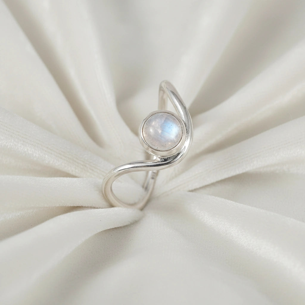 A silver moonstone ring featuring a round moonstone and a wave ring band design at a side angle over a white fabric background