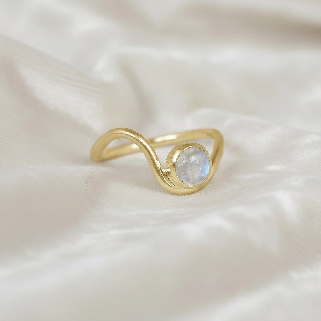 A gold plated moonstone ring featuring a round moonstone and a wave ring band design at a side angle at a white fabric background
