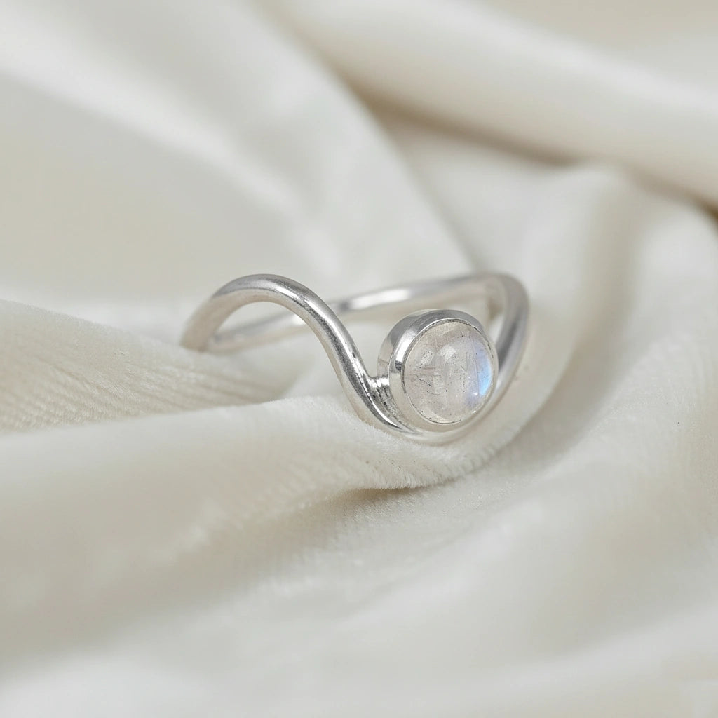 A silver moonstone ring with a round moonstone and a wavy wave ring band at a tilted angle over an off white fabric background