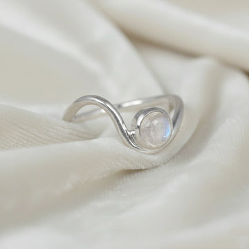 A silver moonstone ring with a round moonstone and a wavy wave ring band at a tilted angle over an off white fabric background