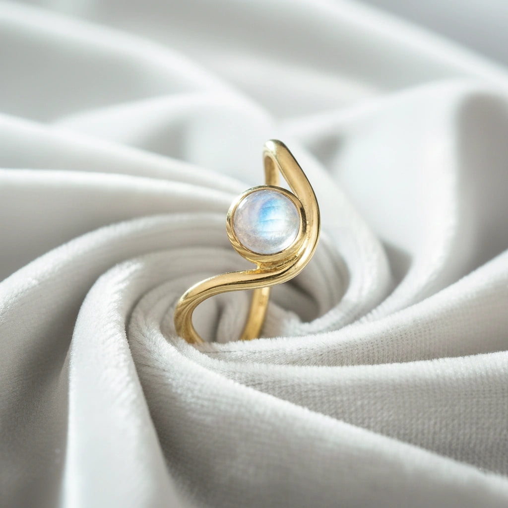 A gold plated moonstone ring with a round moonstone and a wave ring band at a tilted angle at an off white fabric background