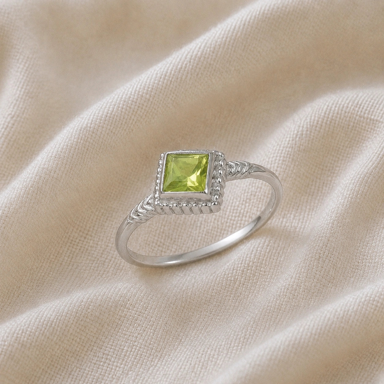 Princess_cut_ring_sterling_silver_with_square_peridot_and_detailed_band_slightly_tilted_view_on_soft_fabric_background