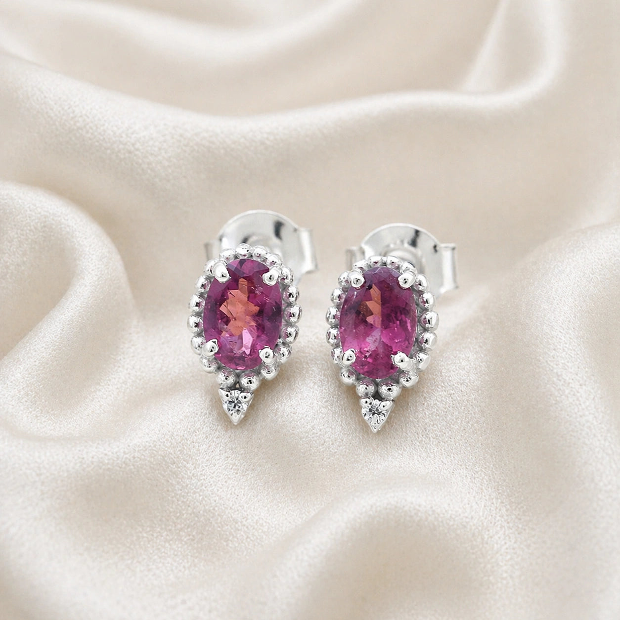 Pink_tourmaline_earrings_in_sterling_silver_with_oval_stone_and_a_white_topaz_shown_front_angle_view_on_soft_fabric_background
