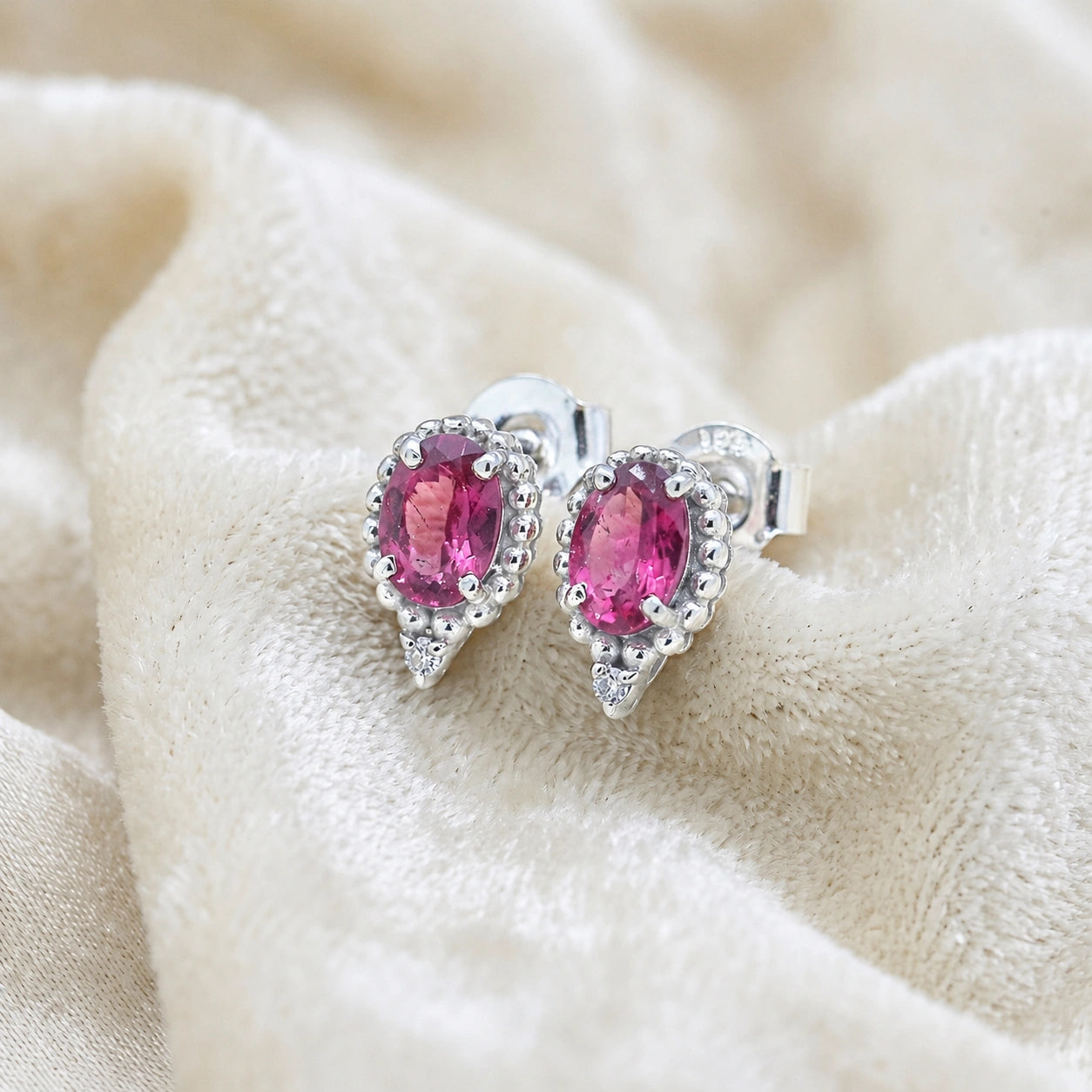Pink_tourmaline_earrings_in_sterling_silver_with_oval_stone_and_a_white_topaz_in_slightly_angle_view_on_soft_fabric_background