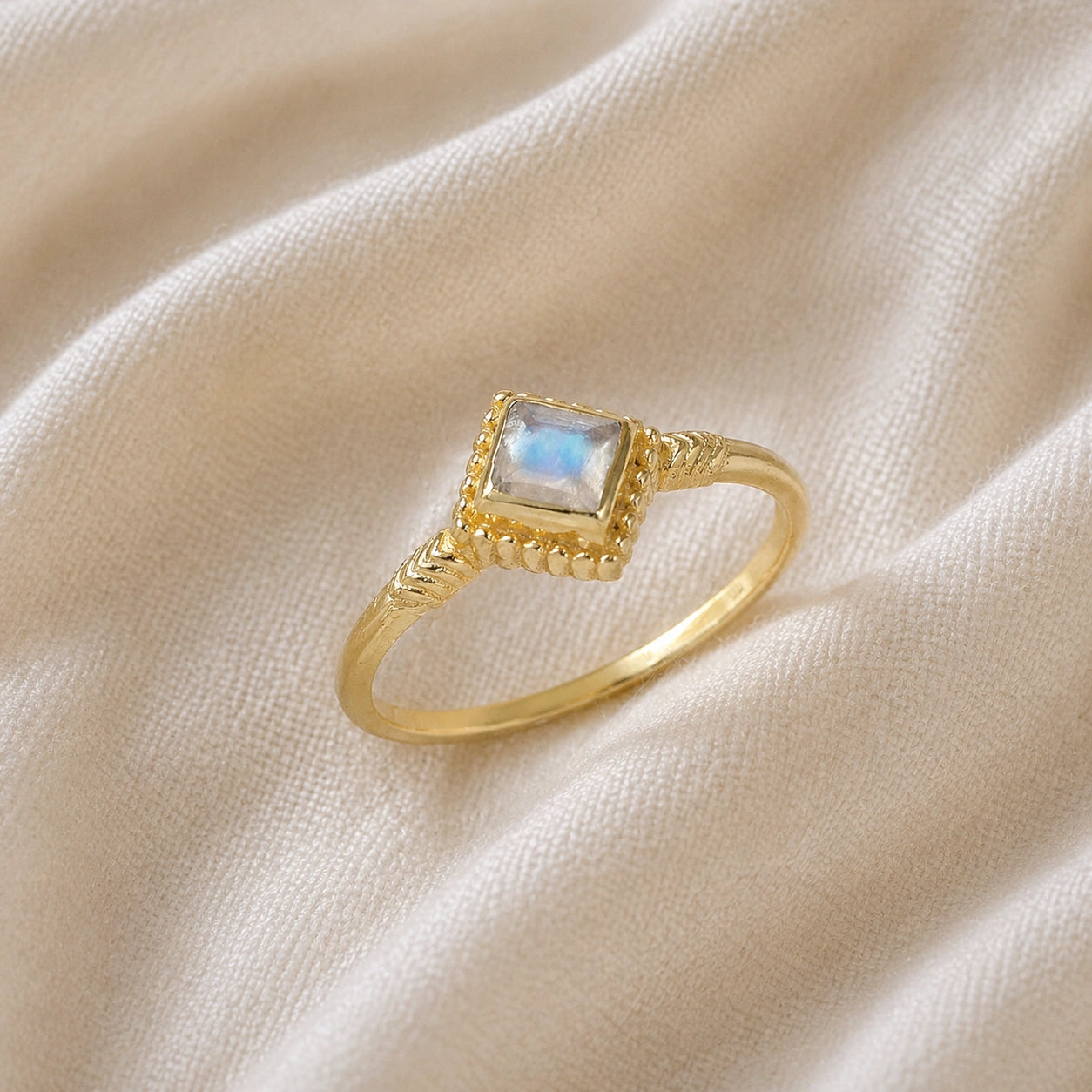 Princess_cut_ring_gold_plated_silver_with_square_moonstone_and_detailed_band_slightly_tilted_view_on_soft_fabric_background