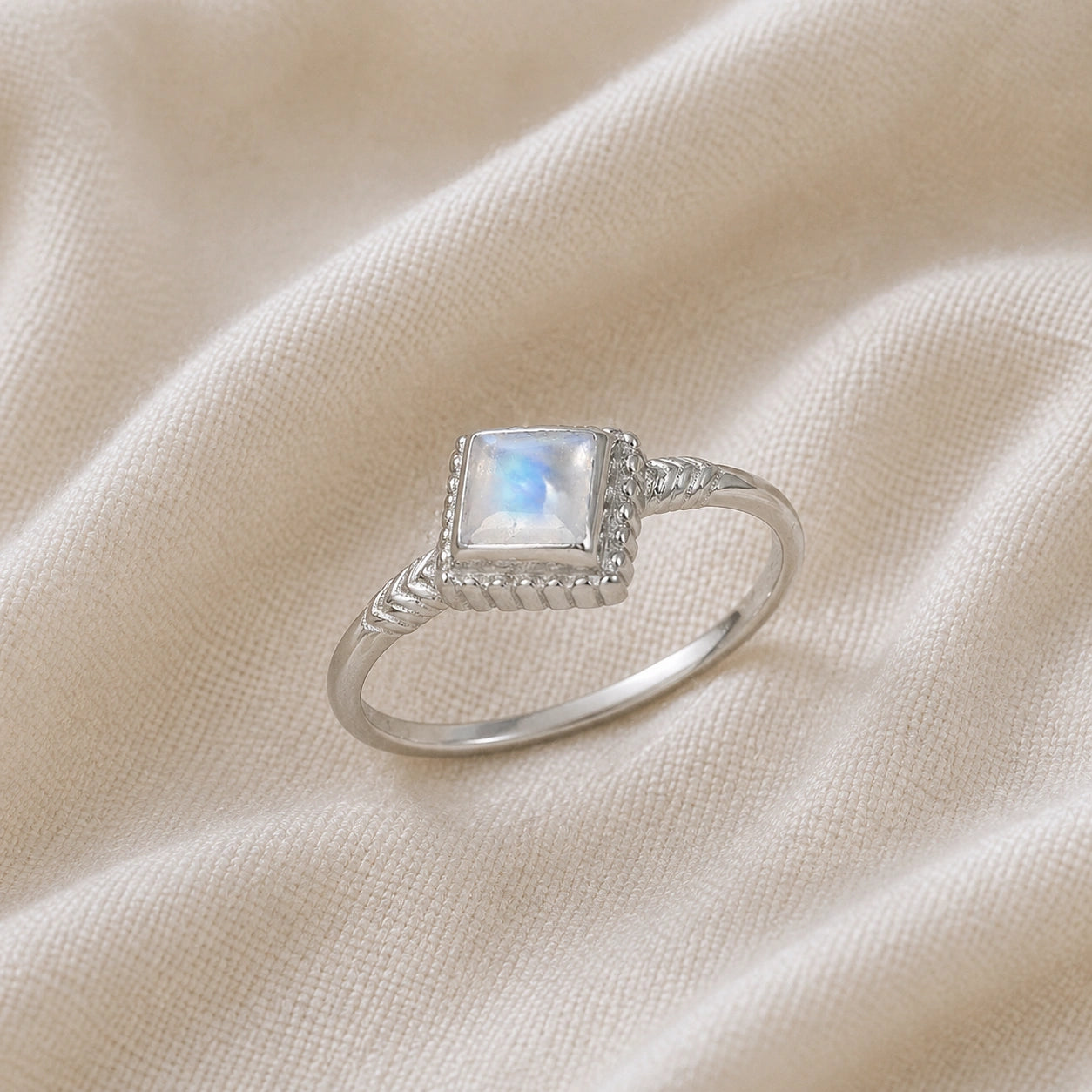 Princess_cut_ring_sterling_silver_with_square_moonstone_and_detailed_band_slightly_tilted_view_on_soft_fabric_background