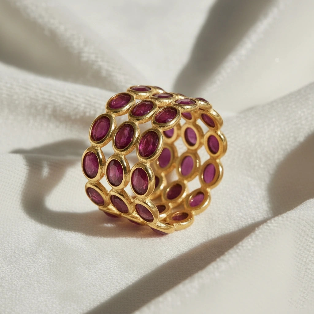 Wide gold ruby eternity ring band featuring three tiers of dark oval rubies mounted on a messy white cloth with an angled view.