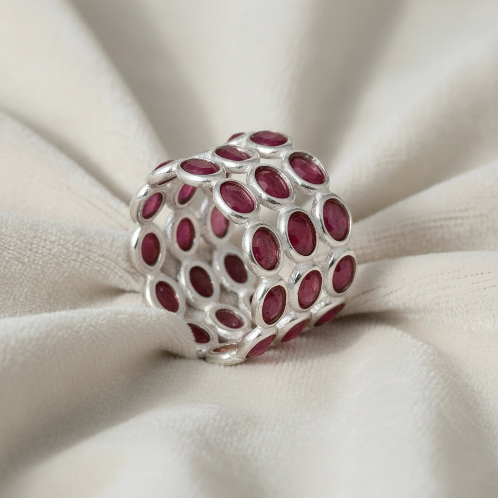 Wide silver ruby band ring featuring three tiers of dark oval rubies mounted on textured white cloth with side profile.