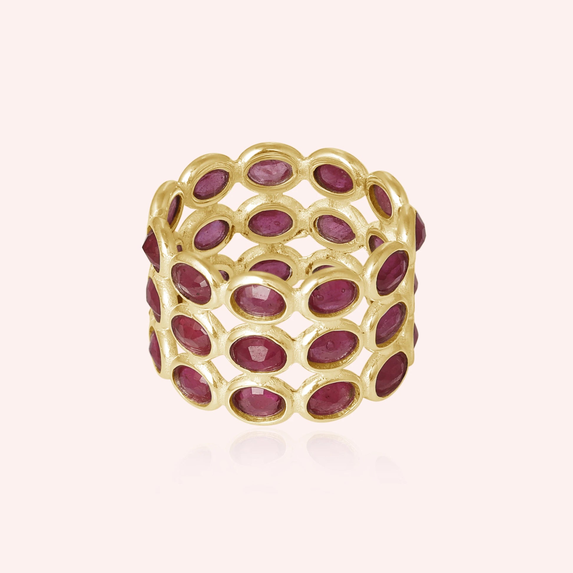 Wide gold ruby eternity ring band featuring oval-cut rubies in an open setting on a reflective pink background from a slight angle.
