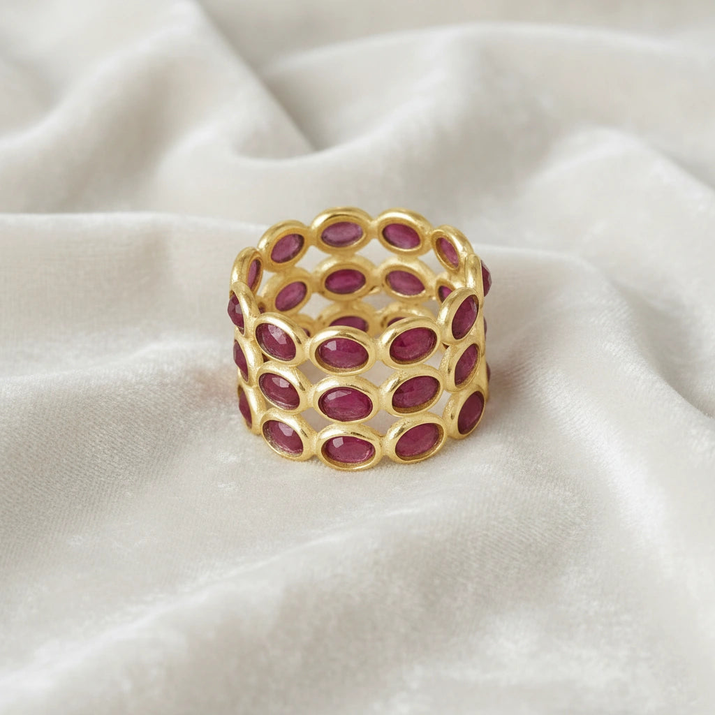 Wide gold ruby eternity ring band featuring three rows of oval rubies mounted on messy white velvet fabric with a front view.
