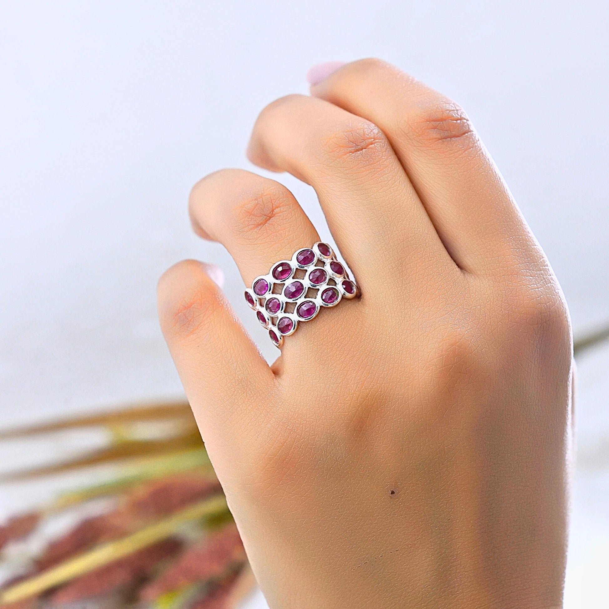 Silver ruby eternity ring band on a woman's finger featuring oval rubies against a light background with flowers at a slight angle.