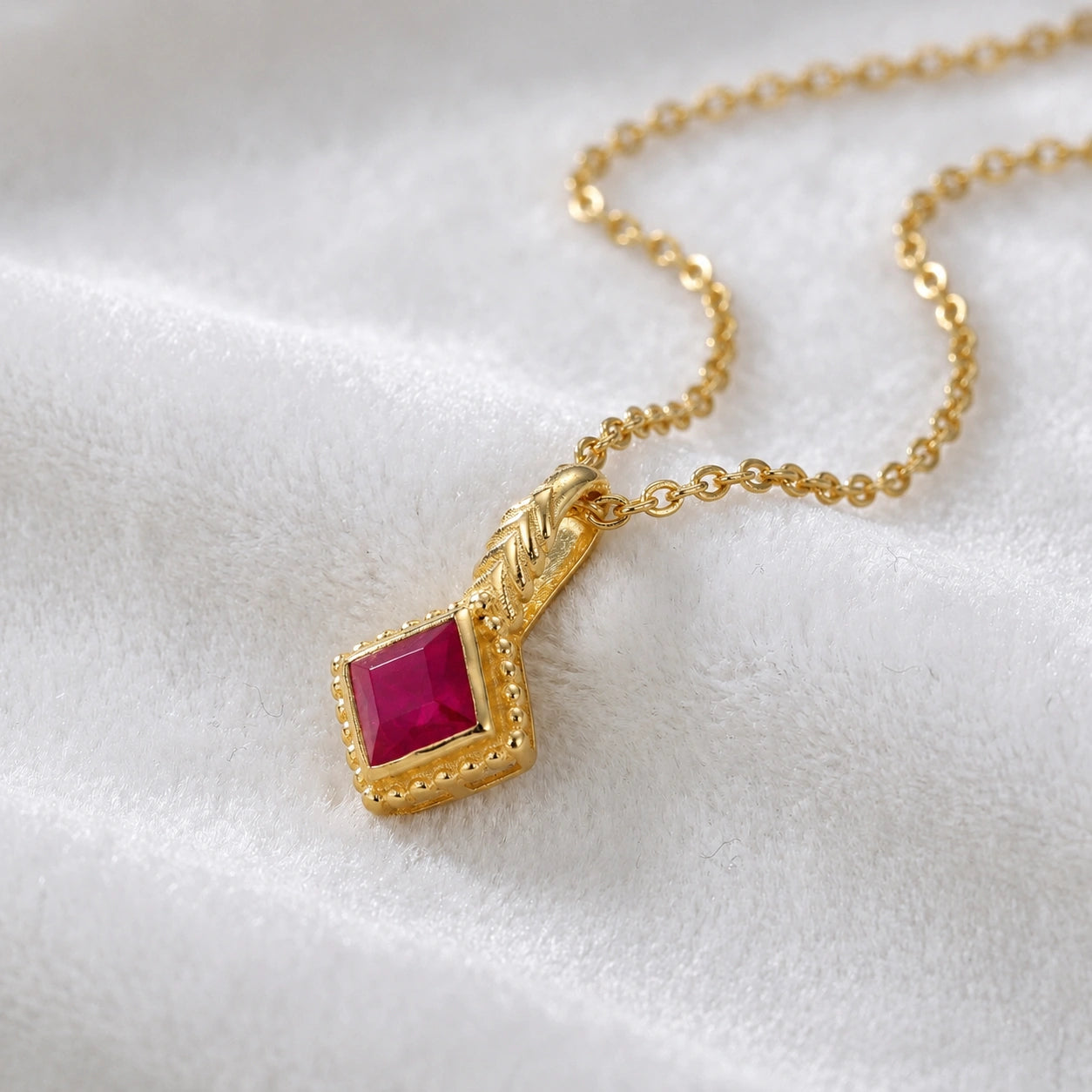 Birthstone_necklace_in_gold_plated_silver_with_square_cut_ruby_pendant_in_slightly_angled_view_on_soft_white_fabric_background