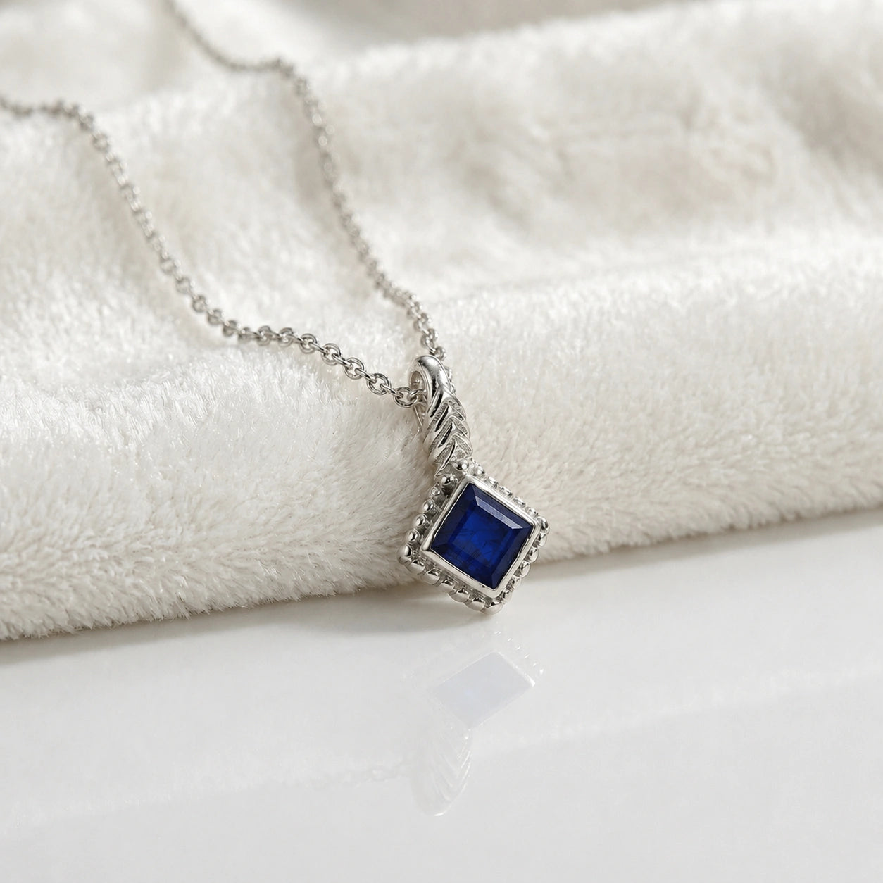 Birthstone_necklace_in_sterling_silver_with_square_cut_sapphire_pendant_in_slightly_angled_view_on_soft_white_fabric_background