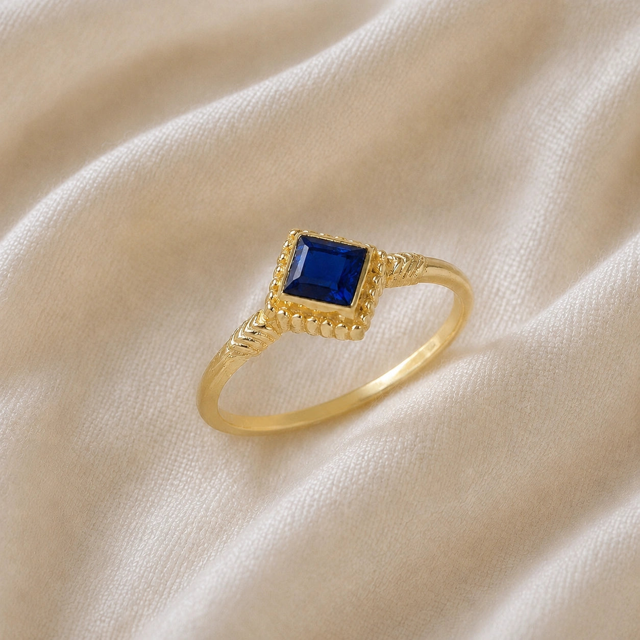 Princess_cut_ring_gold_plated_silver_with_square_sapphire_and_detailed_band_slightly_tilted_view_on_soft_fabric_background