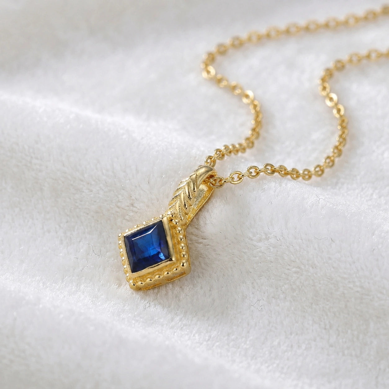 Birthstone_necklace_in_gold_plated_silver_with_square_cut_sapphire_pendant_in_slightly_angled_view_on_soft_white_fabric_background